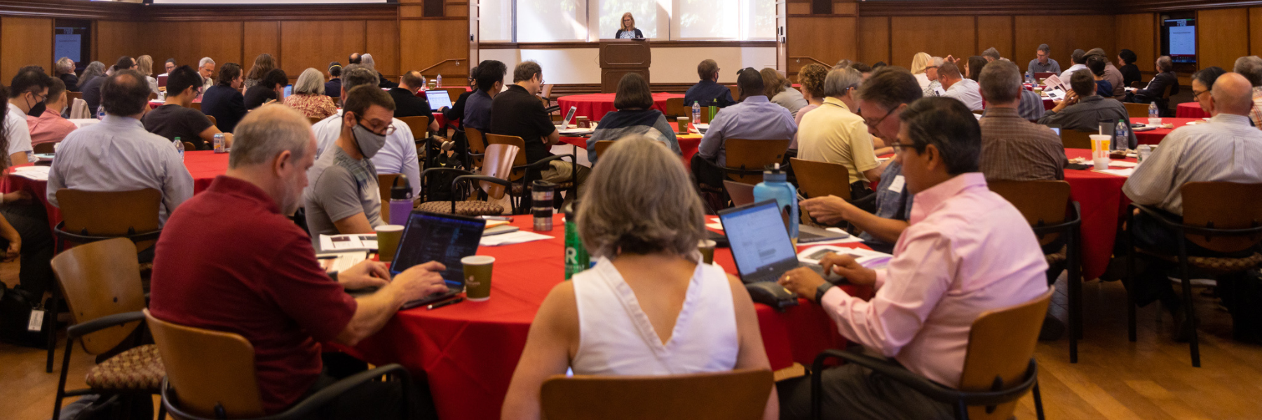 Groups of faculty sit at red, round tables looking through papers or at their laptops. The groups are turned towards a stage in the background of the image, where a blonde woman is speaking at a podium. She is centered on the stage with two screens presenting slides of information.