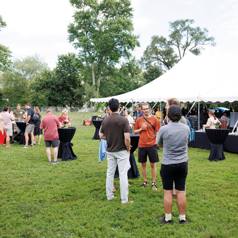 Small groups of people stand and talk on a grassy lawn near a large white event tent, with cocktail tables scattered around.
