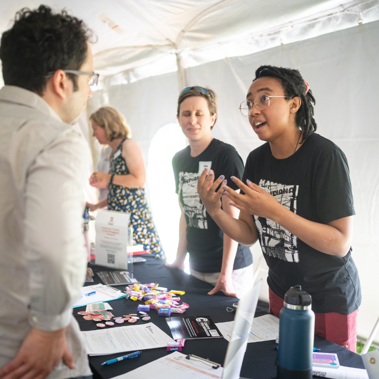 In this image, a woman is standing at a welcome table during the Indiana University New Faculty Orientation Picnic and Resource Showcase. She is engaged in conversation with a faculty member, providing information and assistance. The woman appears attentive and friendly, embodying the welcoming spirit of the event. 