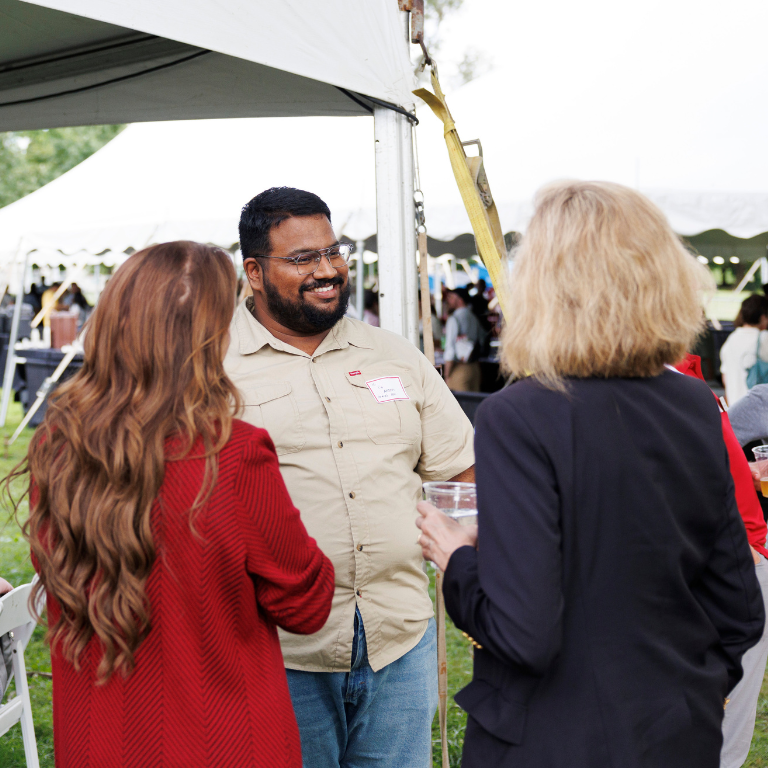 Three people stand and talk under a tent, smiling as they converse during the outdoor event.