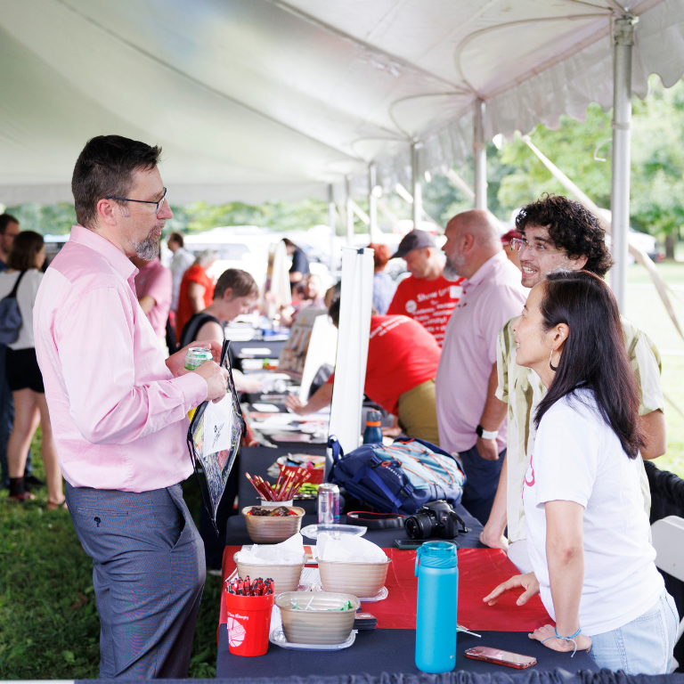 People stand on either side of tables lined with informational materials under a white event tent, talking and smiling.
