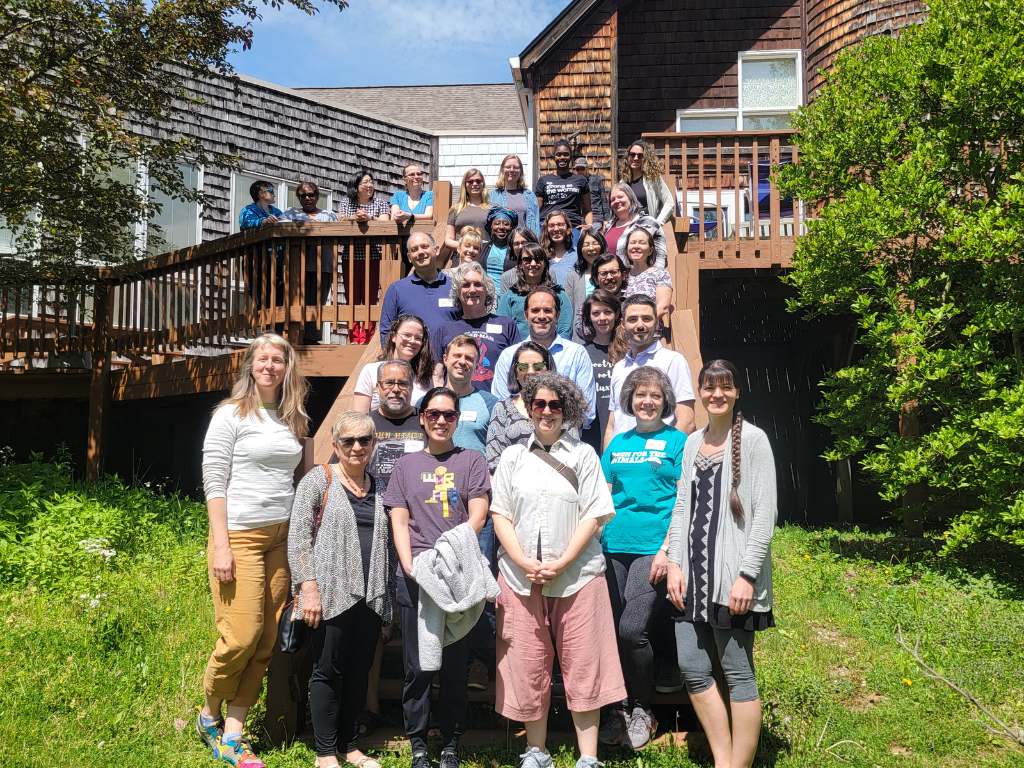 A group of 31 people stands shoulder to shoulder on a wooden staircase facing the camera for the annual Scholarly Writing Retreat. It's a sunny day and everyone has big smiles on their faces. 