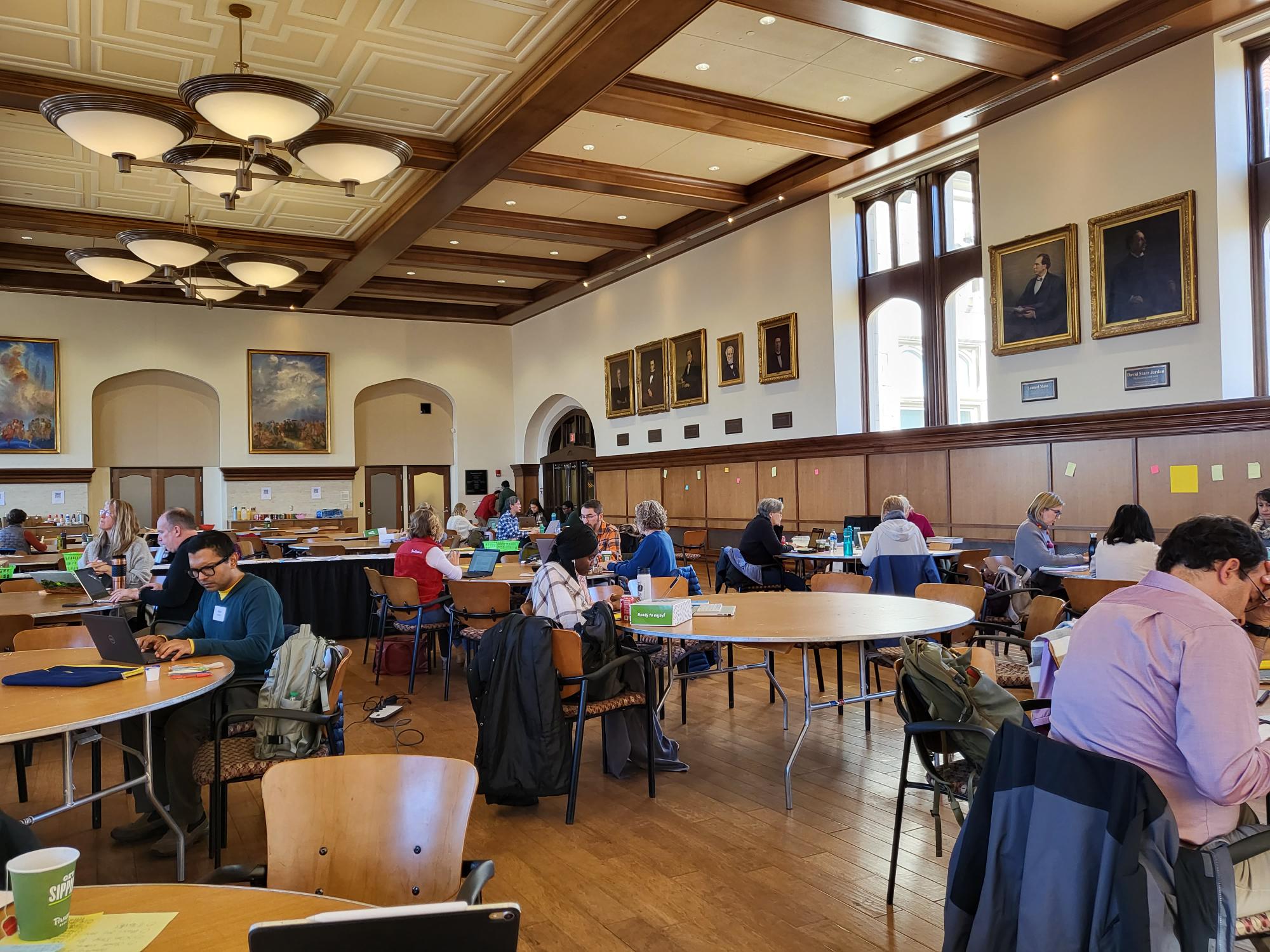 At Woodburn, several groups of people sit together at the tables in the hall. These writing groups all seem to be working on their laptops that are open in front of them. Several members are looking off into the distance, seemingly deep in thought. 