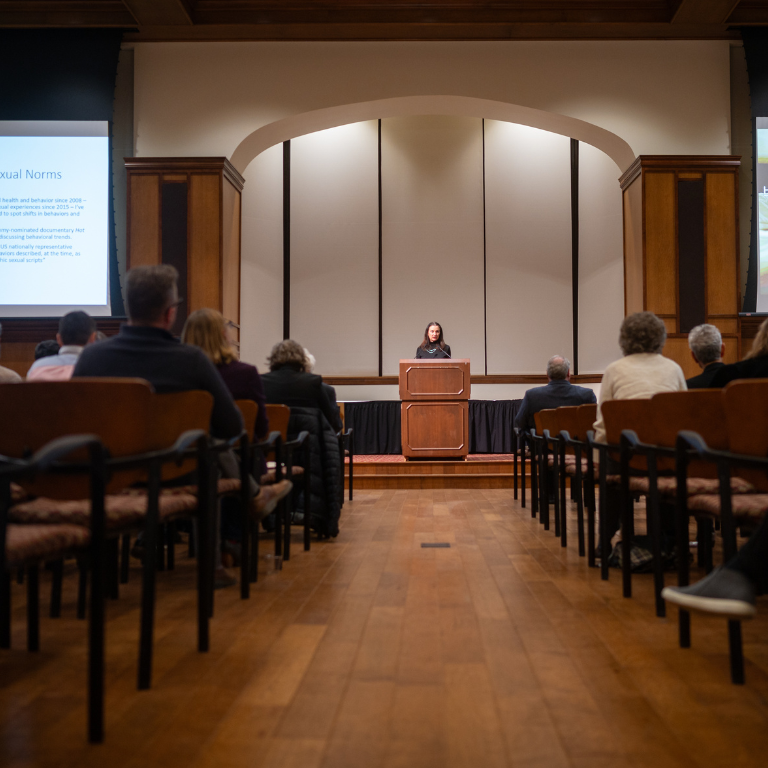 A lecturer stands at a podium in front of a seated audience. 